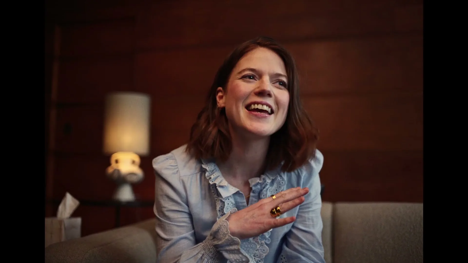A woman with brown hair and a light blue ruffled shirt sits on a couch, laughing and gesturing with her hands in a dimly lit room.
