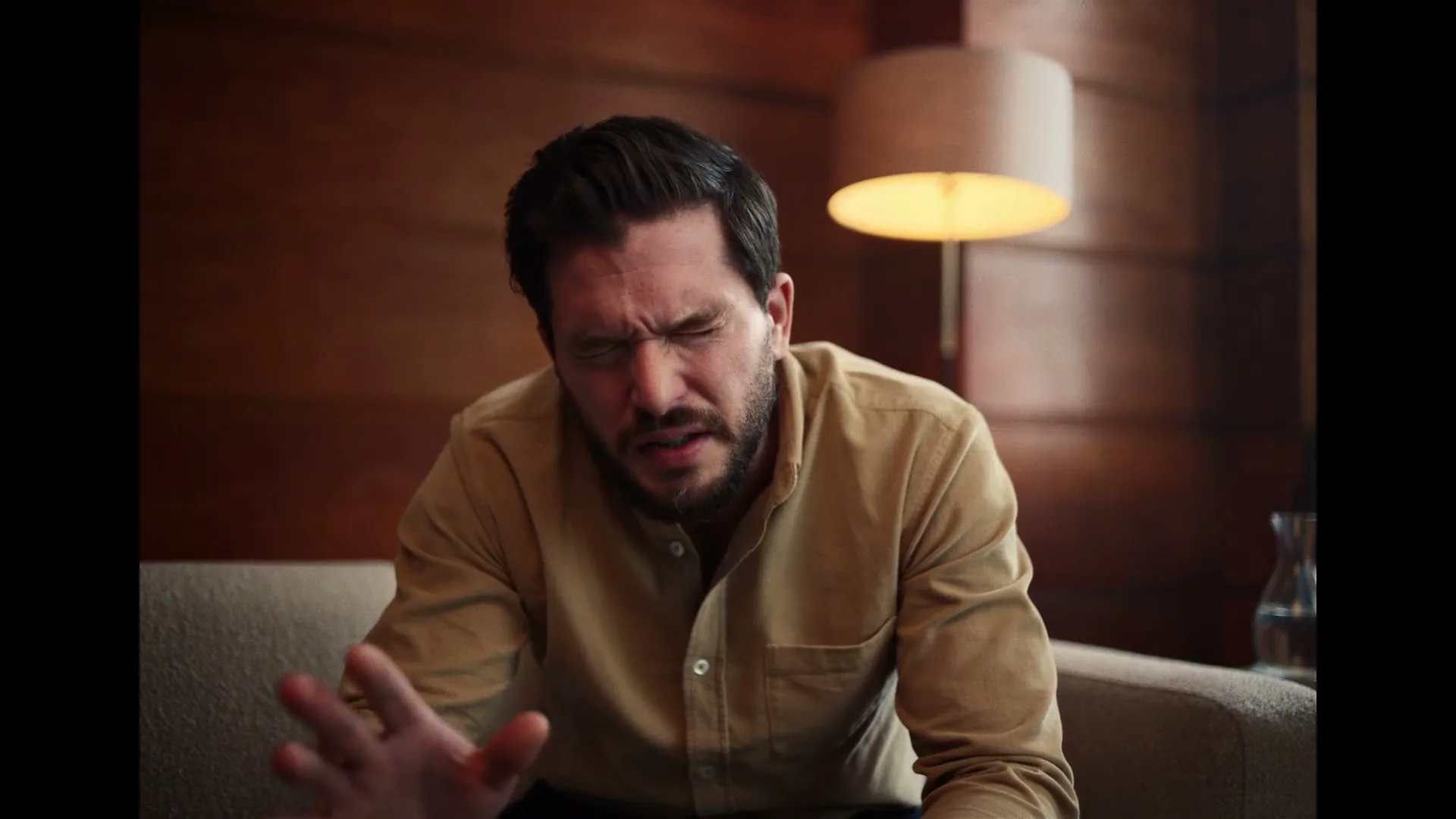 A man with dark hair and a beard, wearing a tan shirt, sits on a couch in a dimly lit room, looking frustrated and gesturing with his hand.