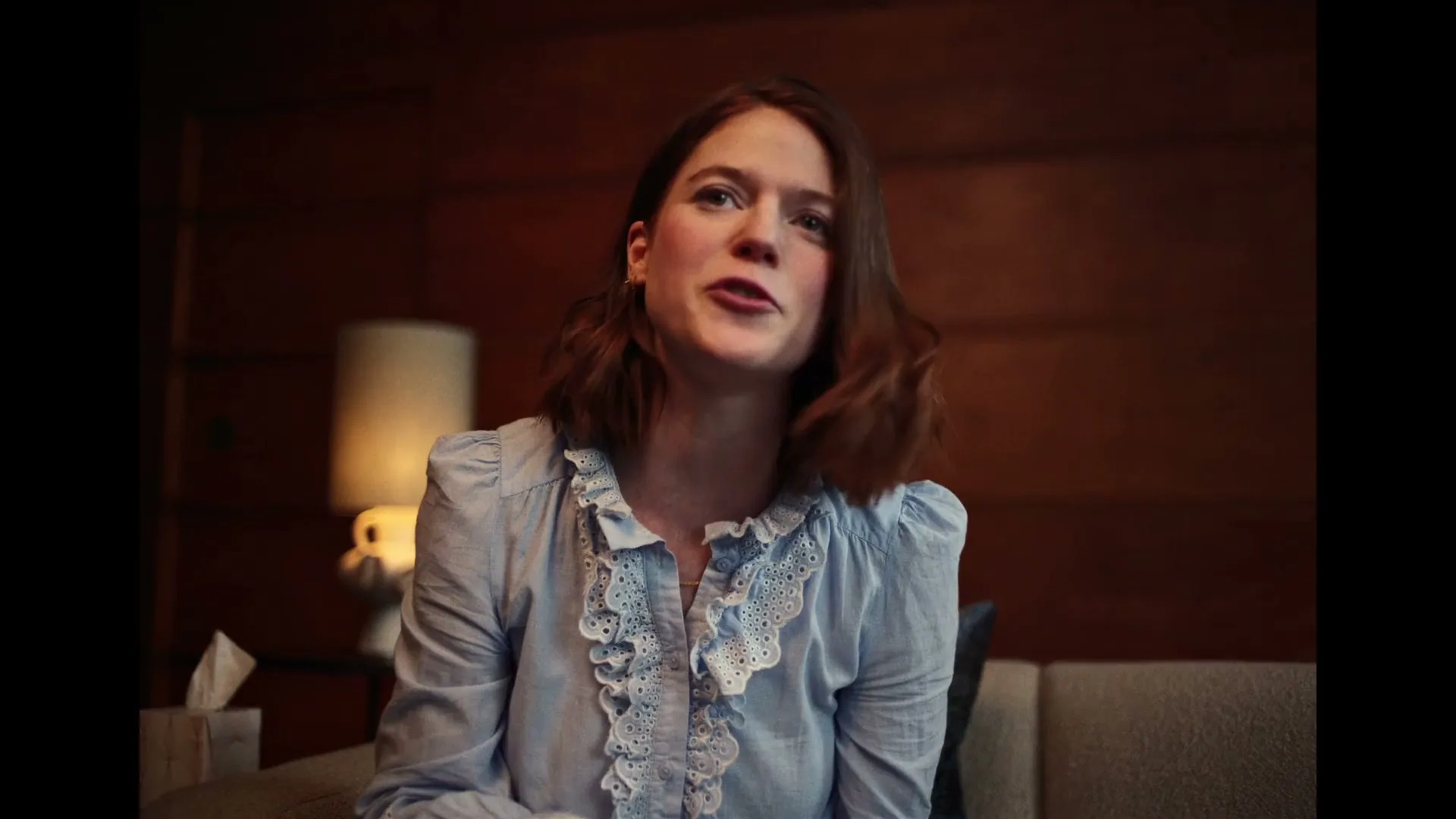 A young woman with brown hair and a blue ruffled shirt sits on a couch, talking animatedly in a dimly lit room with wood paneling.