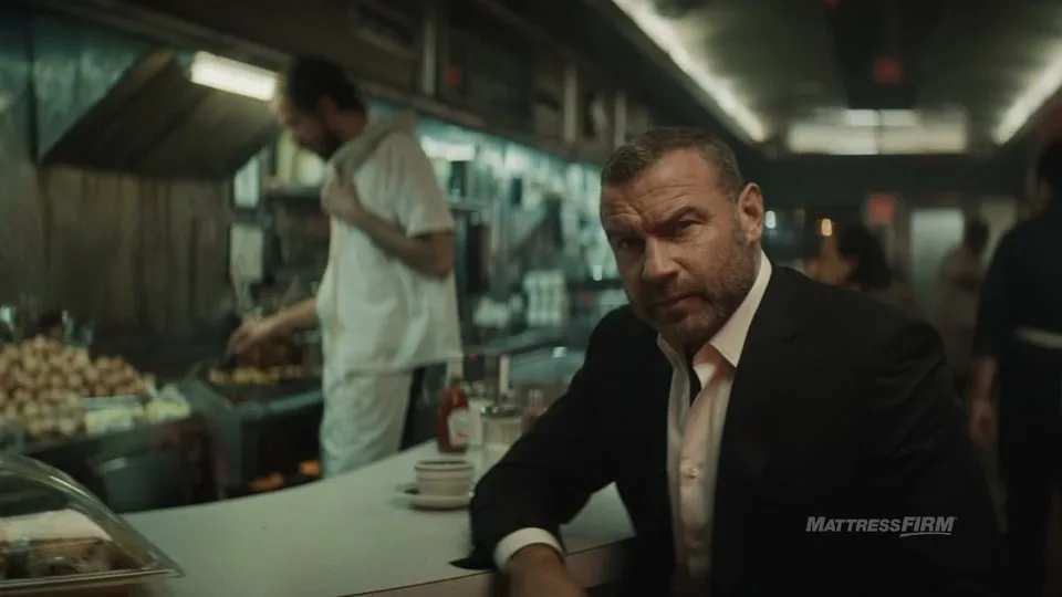 Liev Schreiber, a man in his 50s, sits at a diner counter, looking directly at the camera, while a cook works in the background.