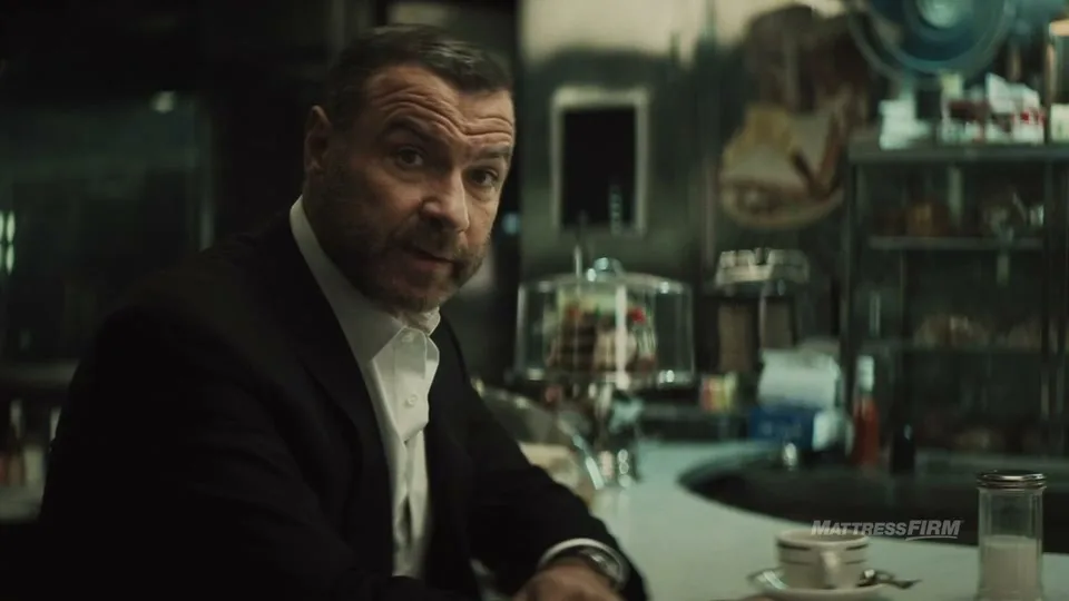A man with a beard in a suit sits at a diner counter, looking directly at the camera with a serious expression.