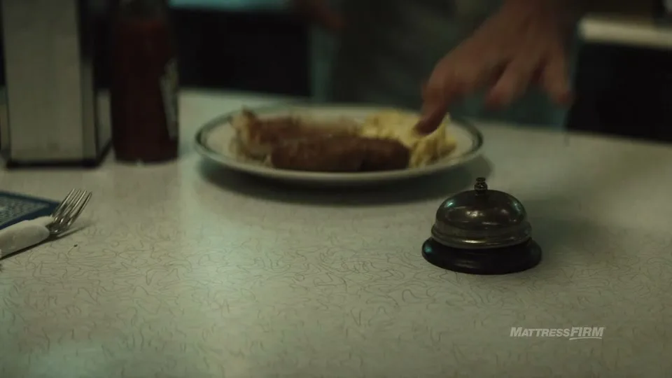 A person's hands are seen in the background as a plate of food is placed on a diner counter next to a service bell.