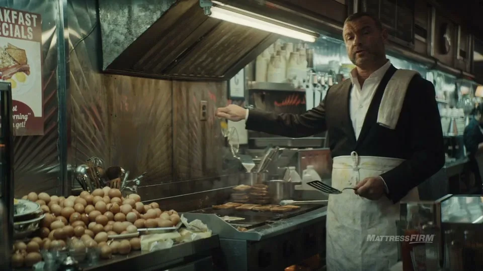 A man in a suit and apron stands behind a diner grill, holding a spatula and gesturing, with a pile of eggs in the foreground.