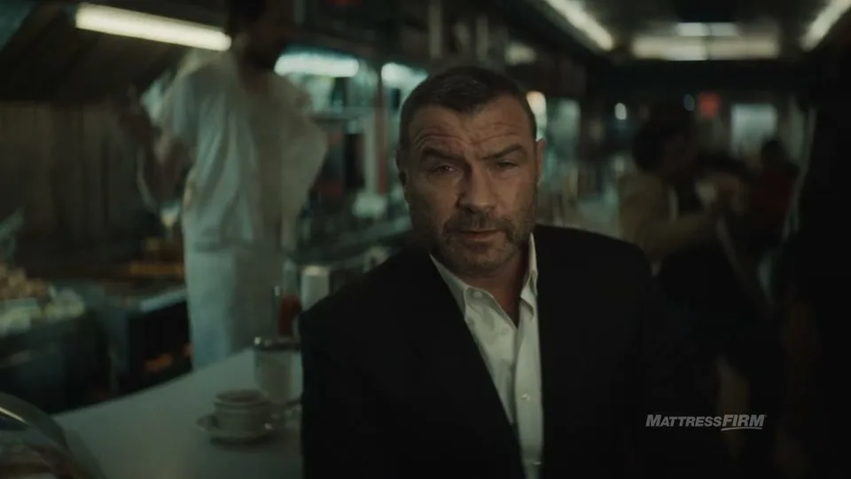 A man in a suit sits at a diner counter, looking directly at the camera, while a chef works in the background.
