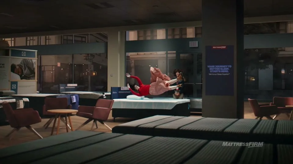 A woman in a red top and pink skirt floats above a mattress in a store, while another woman watches from behind, in a wide shot.
