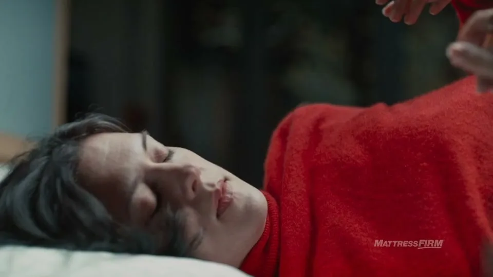 A young woman with dark hair, wearing a red sweater, lies on a bed with her eyes closed, as a hand gently touches her head.