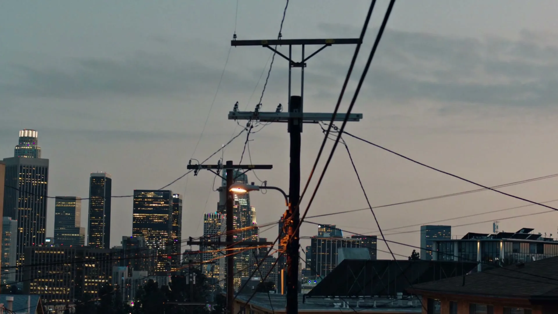 A wide shot of a city skyline at dusk, with power lines and utility poles in the foreground, and buildings with illuminated windows in the background.