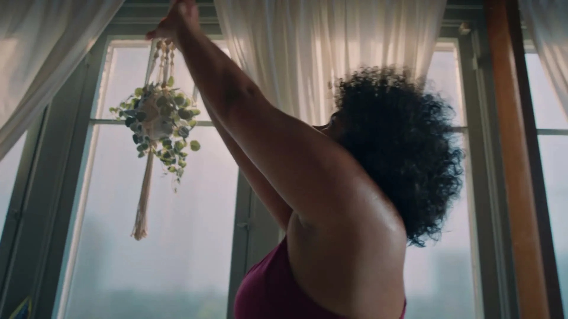 A woman with curly hair in a magenta top stretches her arms upwards in front of a window with white curtains and a hanging plant.