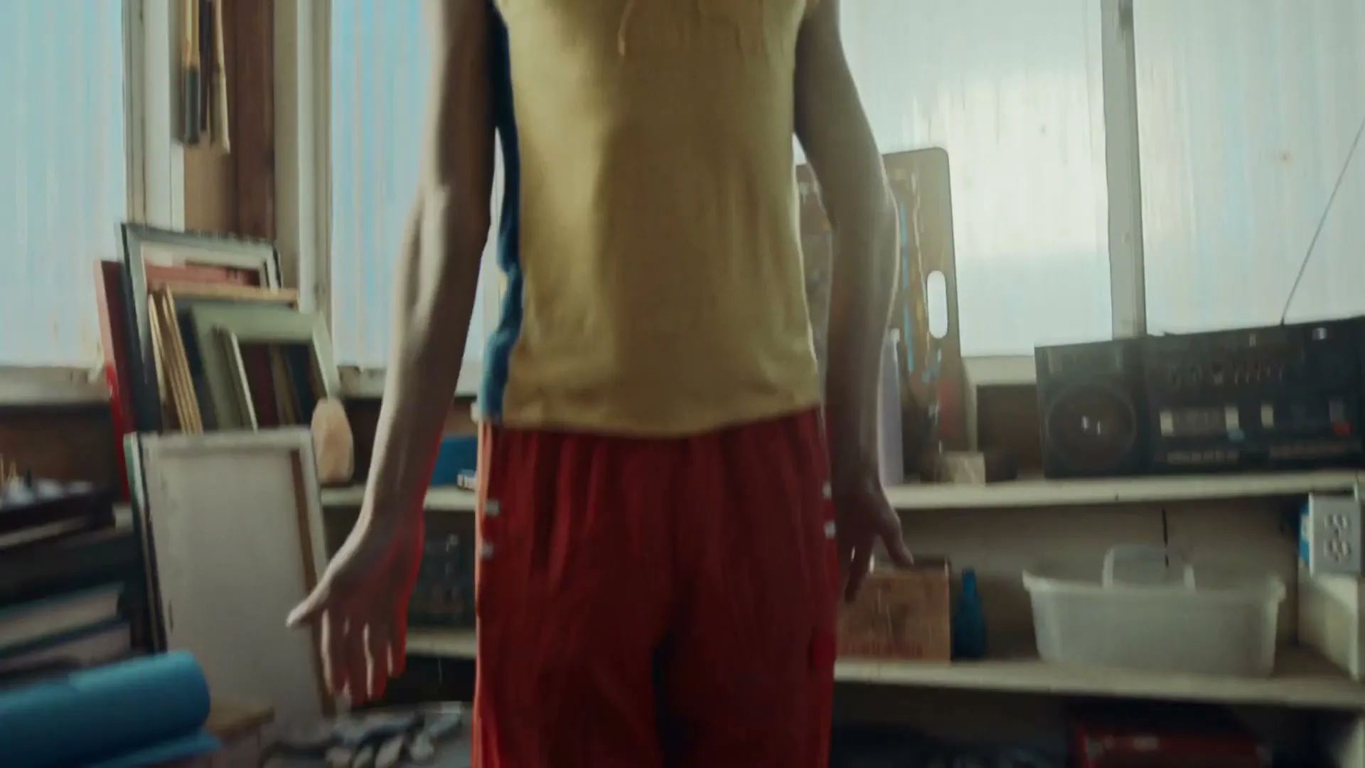 A man with an afro, wearing a yellow tank top and red pants, stretches in a cluttered room with a yoga mat and weights.