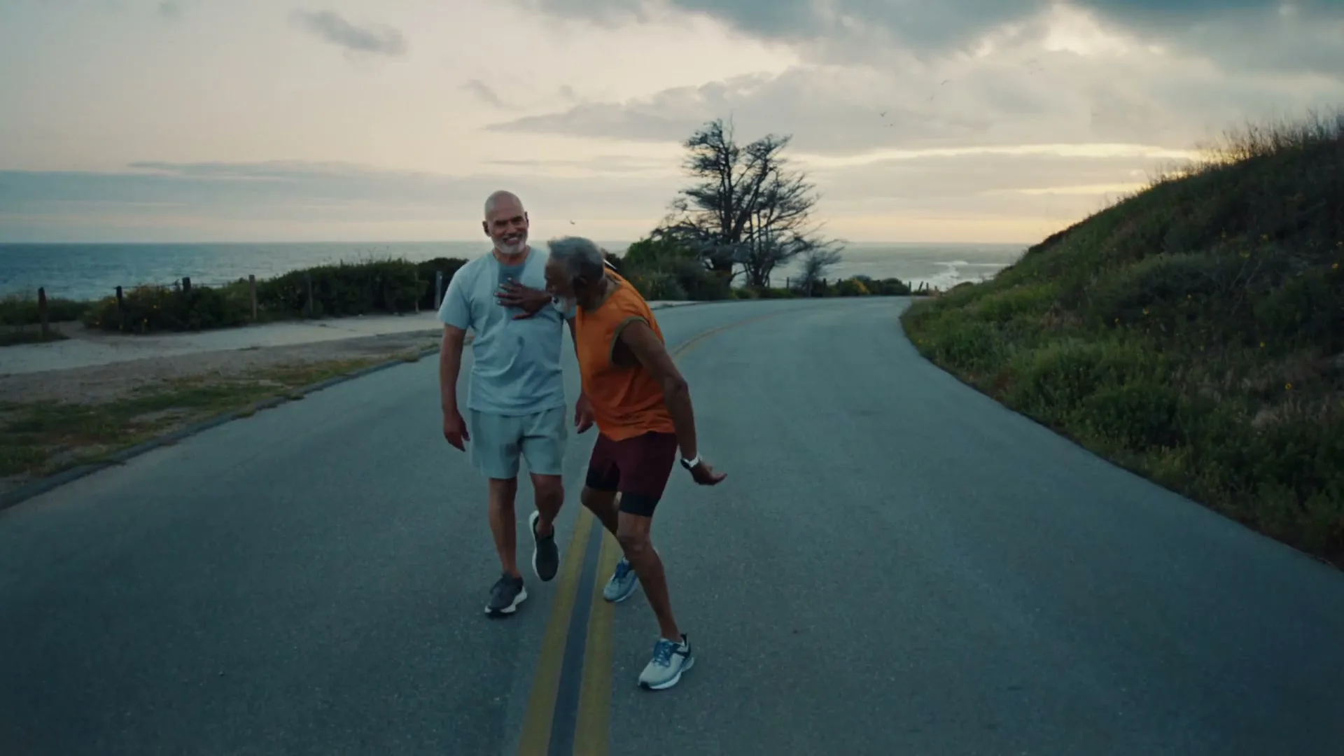 Two older men, one bald and one with gray hair, walk and dance on a coastal road at dusk, with the ocean in the background.