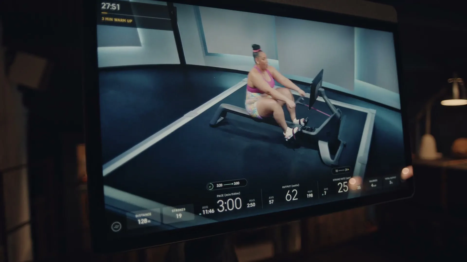 A woman with a high bun and pink sports bra rows on a Peloton machine, displayed on a screen in a dimly lit room.
