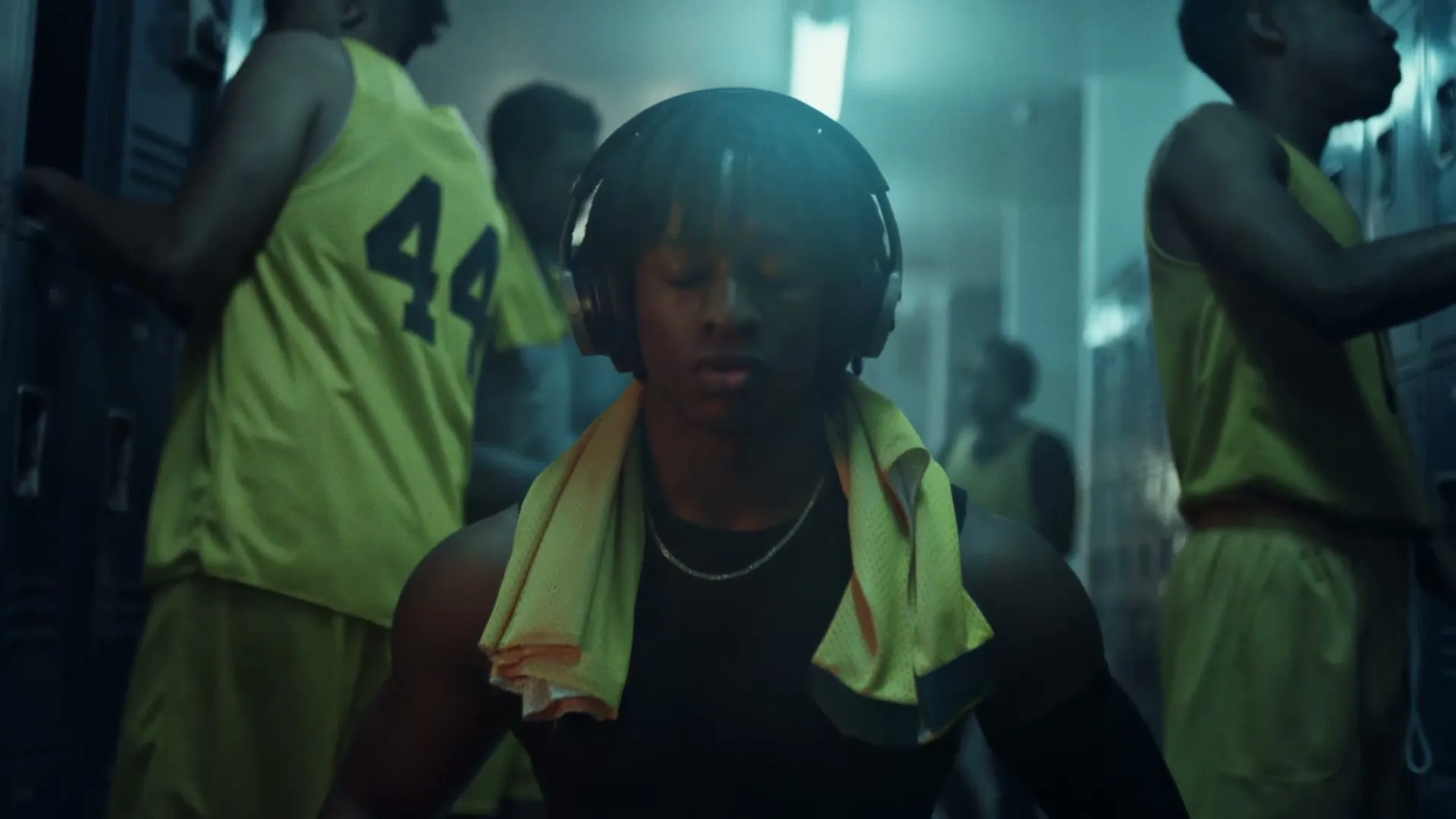 A young Black man with dreadlocks and headphones meditates in a locker room, eyes closed, with a yellow towel around his neck, surrounded by other basketball players.