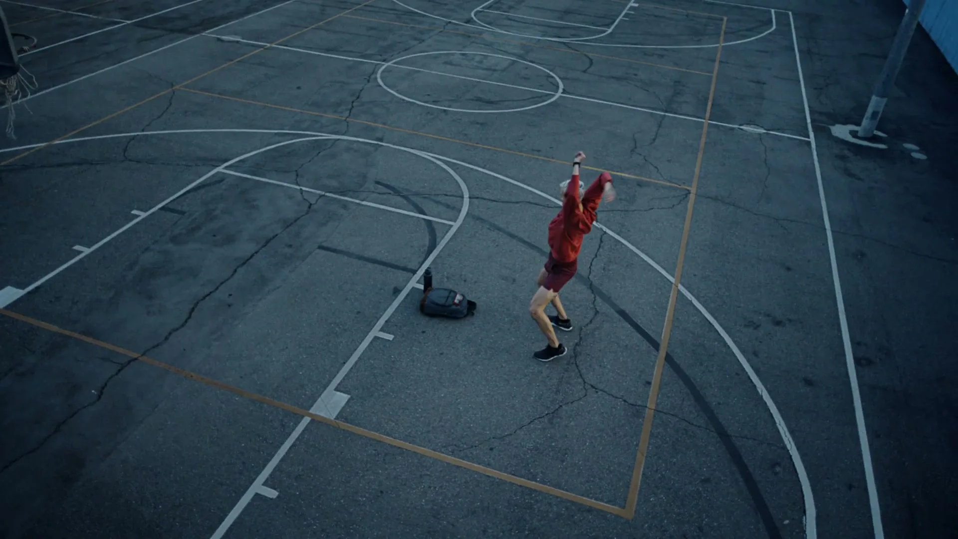 A man with white hair in a red shirt and shorts stretches on an outdoor basketball court with a gym bag and water bottle nearby.
