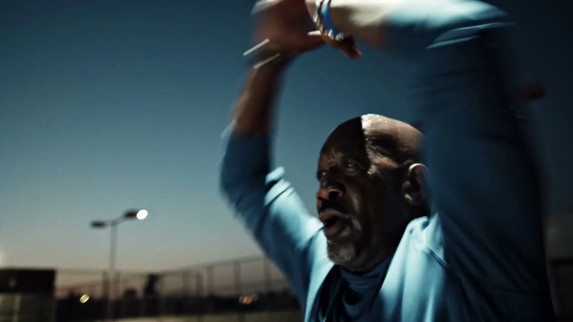 A bald, older man in a blue hoodie exercises with his arms raised on an outdoor court at dusk.