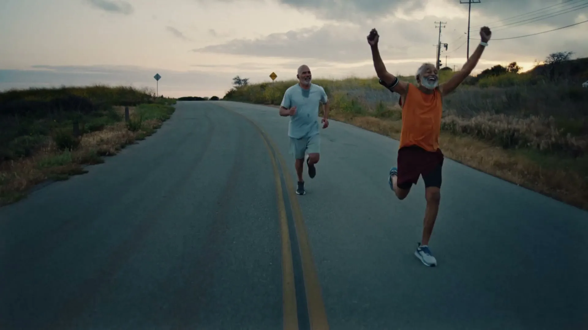 Two older men run on a winding road at dusk, one in an orange shirt raises his arms in triumph, while the other smiles.