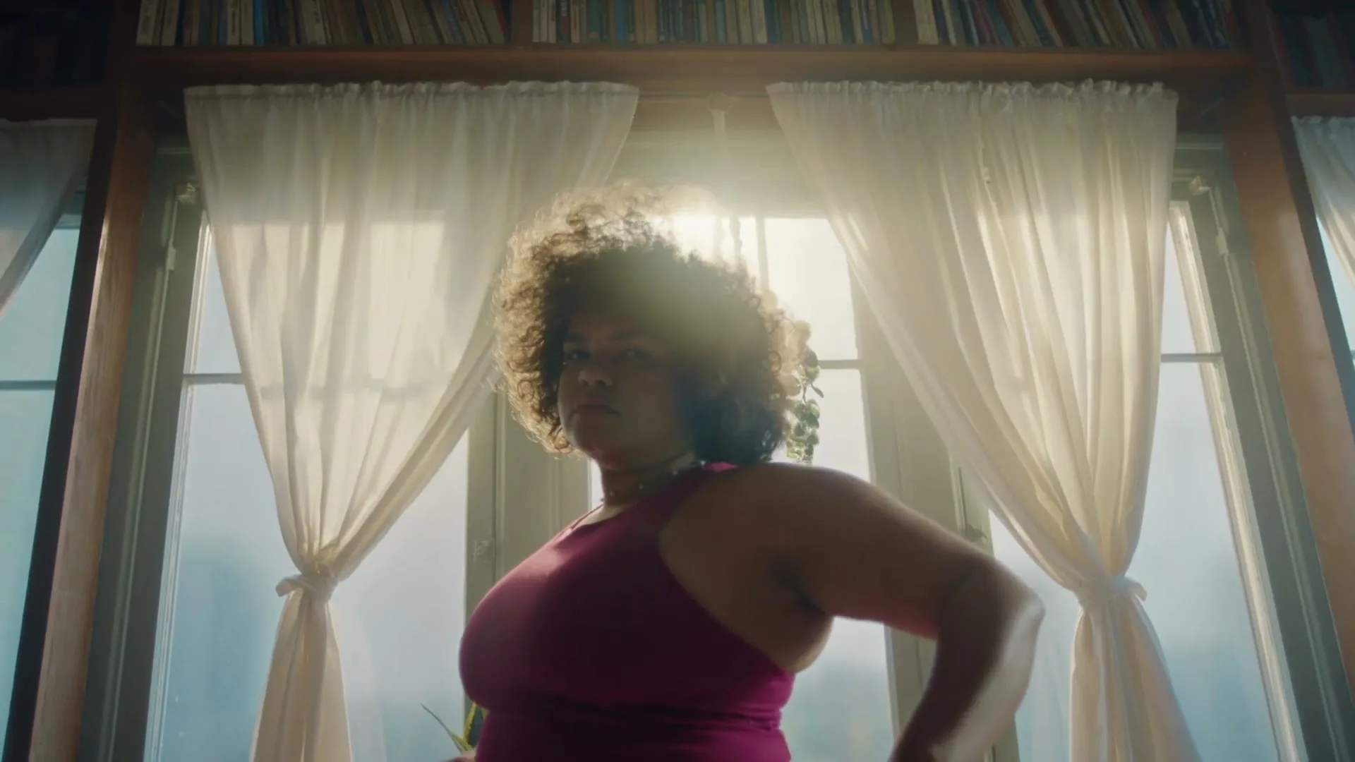 A woman with curly hair, wearing a pink top, stands in front of a sunlit window with white curtains, looking directly at the camera.