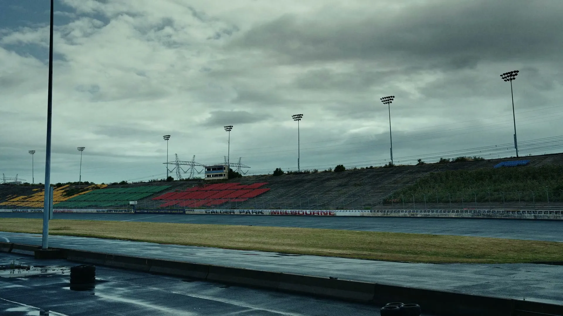 An empty, weathered race track with colorful grandstands and tall light poles under a cloudy sky.