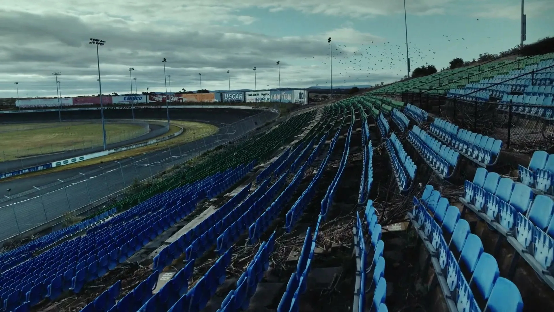 An abandoned race track with empty, dilapidated blue and green stadium seats under a cloudy sky, with birds flying in the distance.