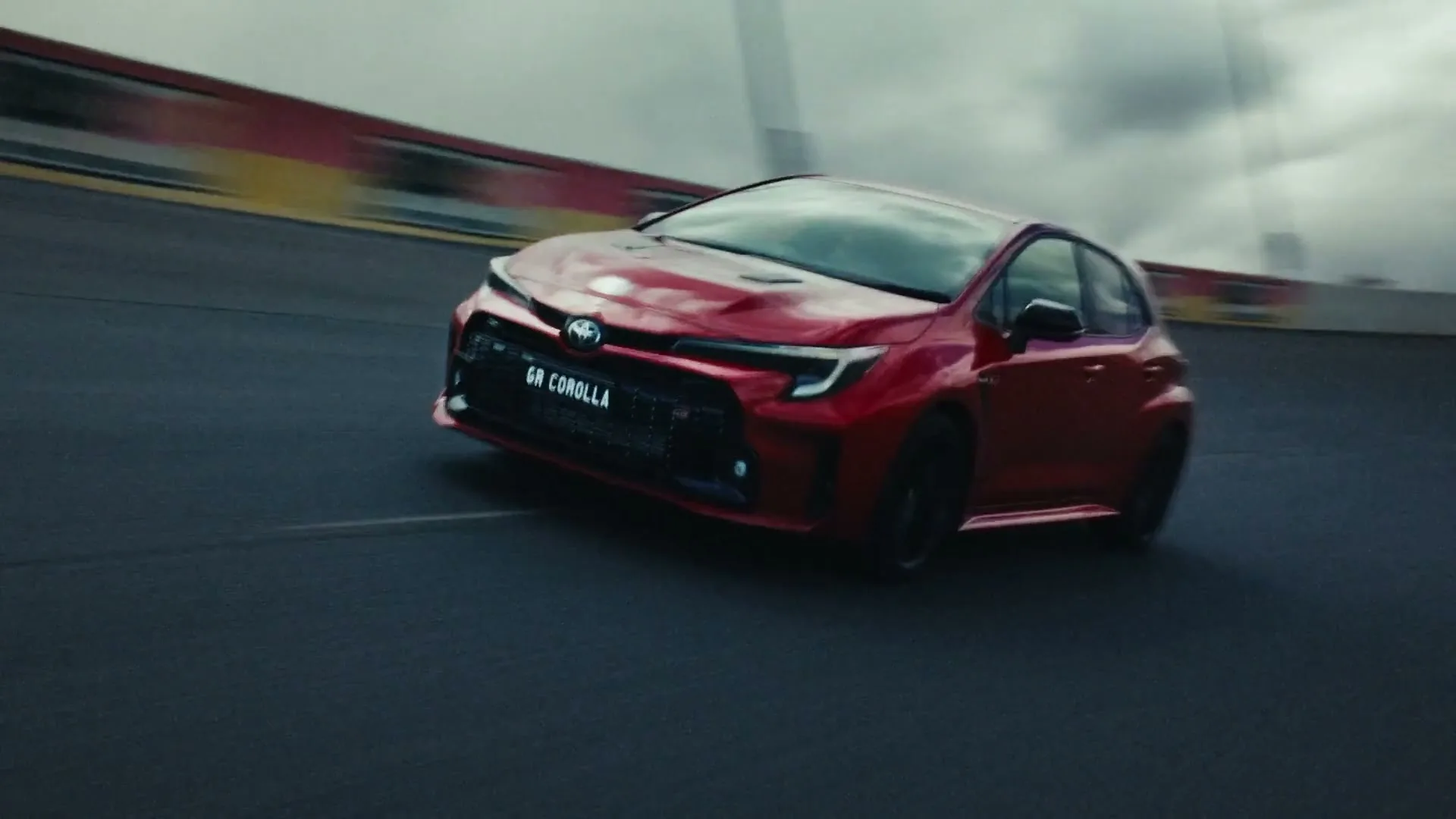 A red Toyota GR Corolla races around a track, with motion blur indicating high speed under a cloudy sky.