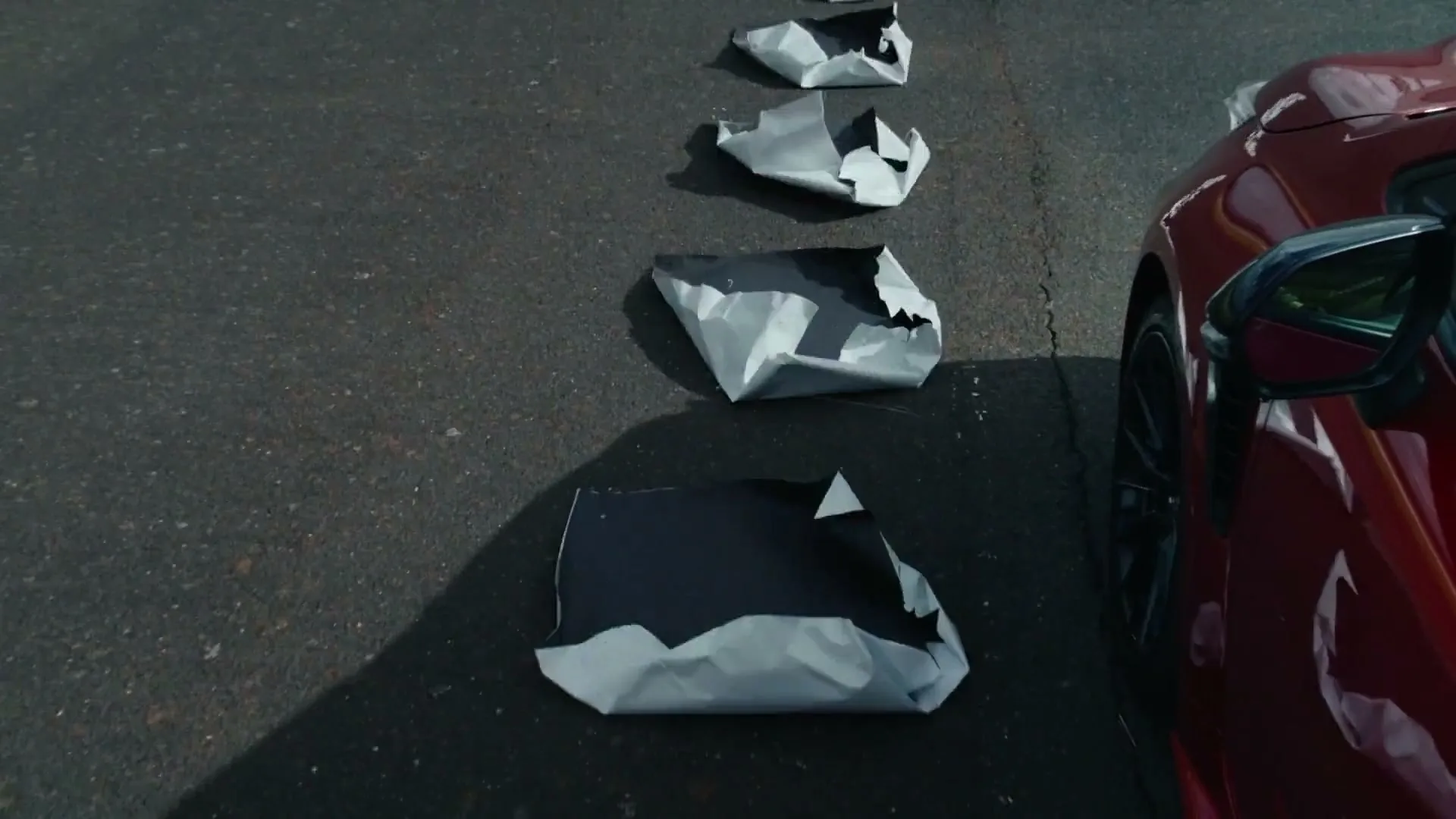 A red sports car is parked next to a line of black squares on the asphalt, which then crumple into silver shapes.