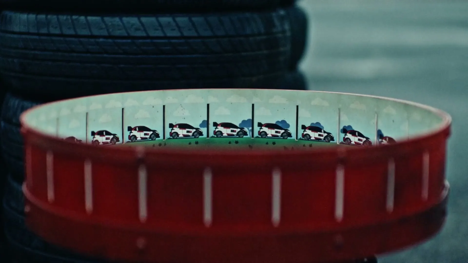 A close-up shot shows a red zoetrope with small, animated race cars inside, spinning rapidly against a blurred background of stacked tires.