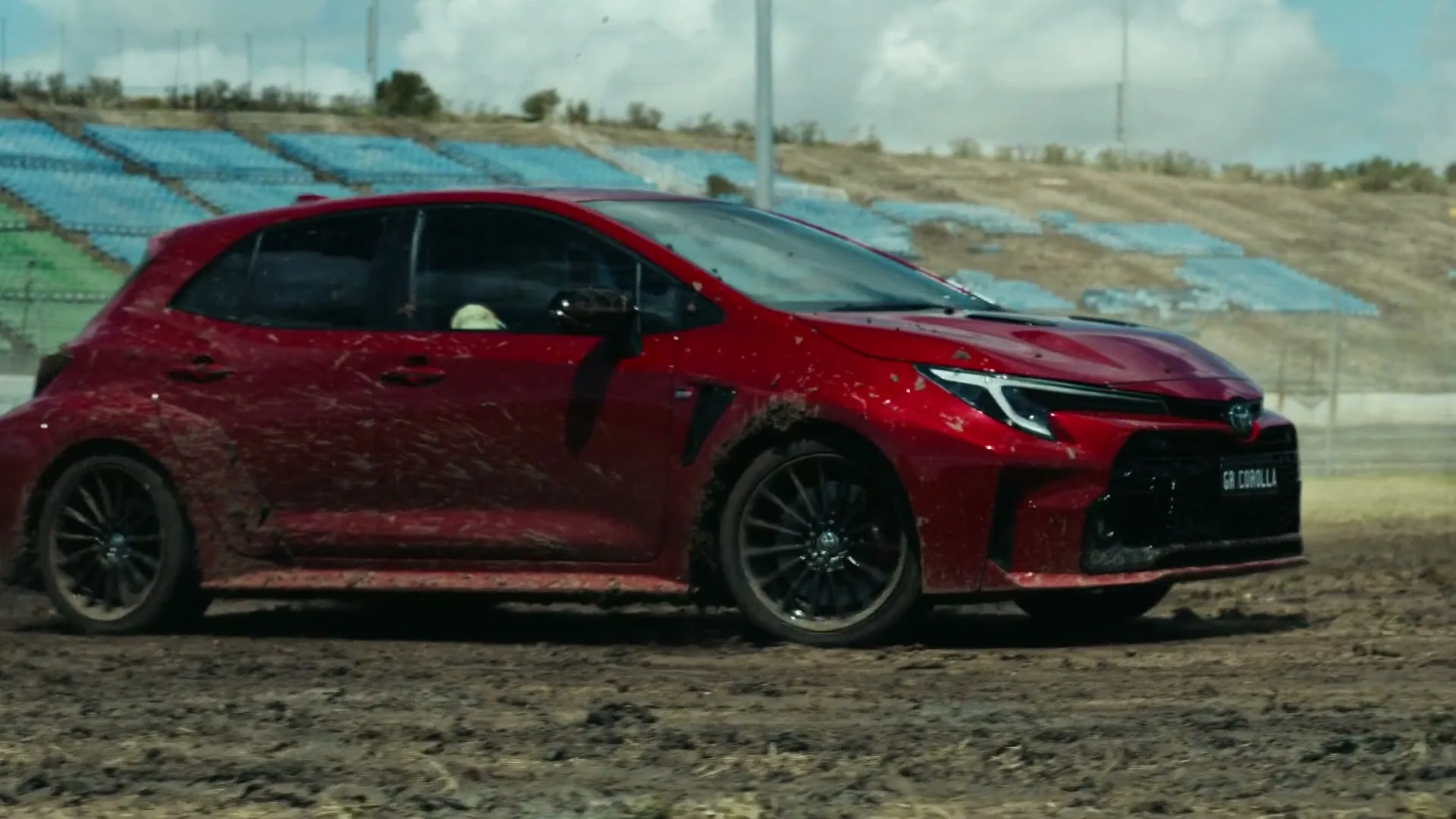 A red Toyota GR Corolla, covered in mud, drifts on a dirt track in front of empty stadium bleachers under a cloudy sky.