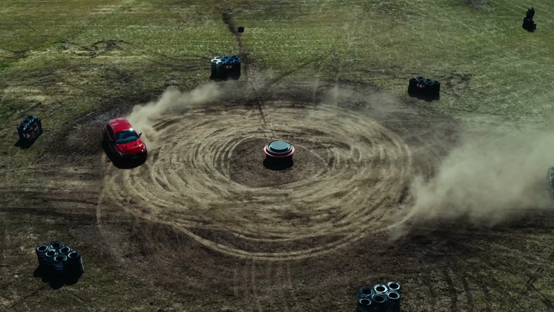 An aerial view shows a red Toyota SUV performing a donut around a central object in a dusty, grassy field, kicking up dirt.