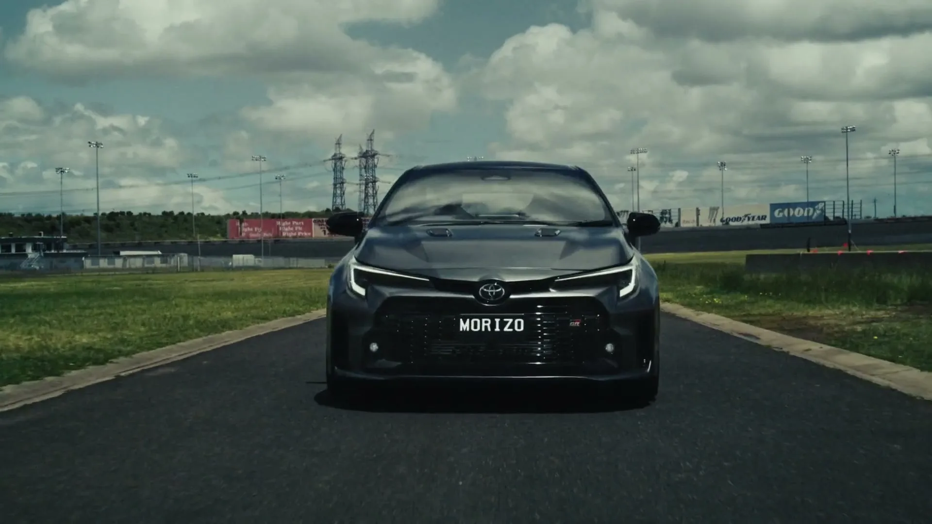 A dark grey Toyota GR Corolla Morizo Edition drives directly towards the camera on a race track under a cloudy sky.