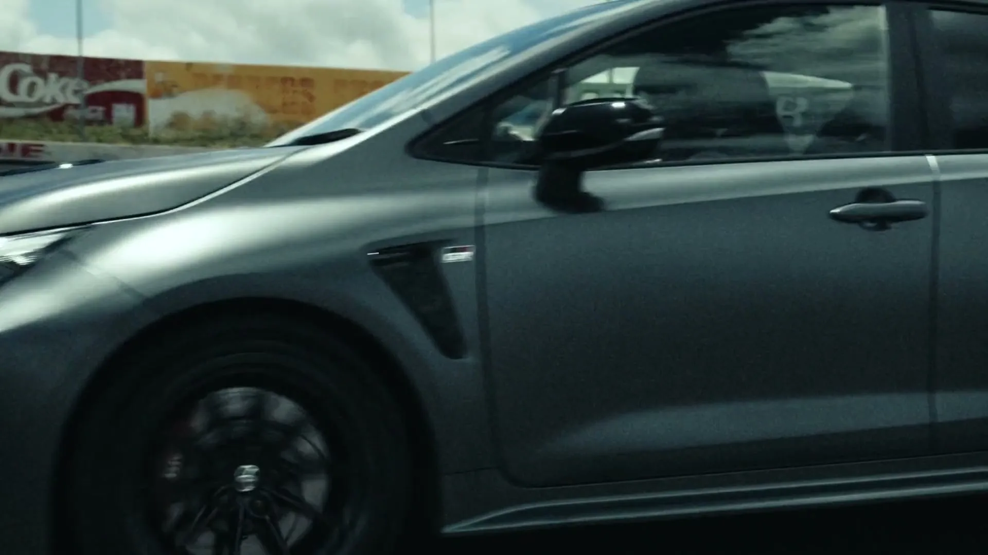 A dark grey Toyota GR Corolla races along a track, with a blurred background of a fence and billboards under a cloudy sky.