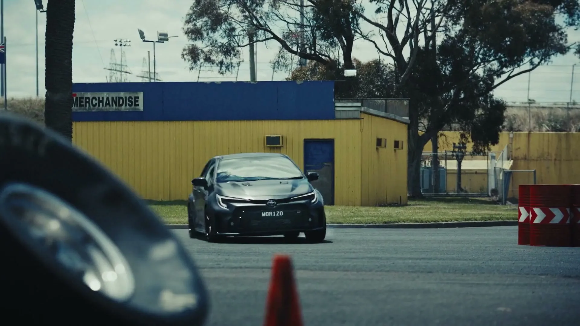 A dark grey Toyota GR Corolla hatchback drifts around a corner on an asphalt track, with a yellow building and trees in the background.