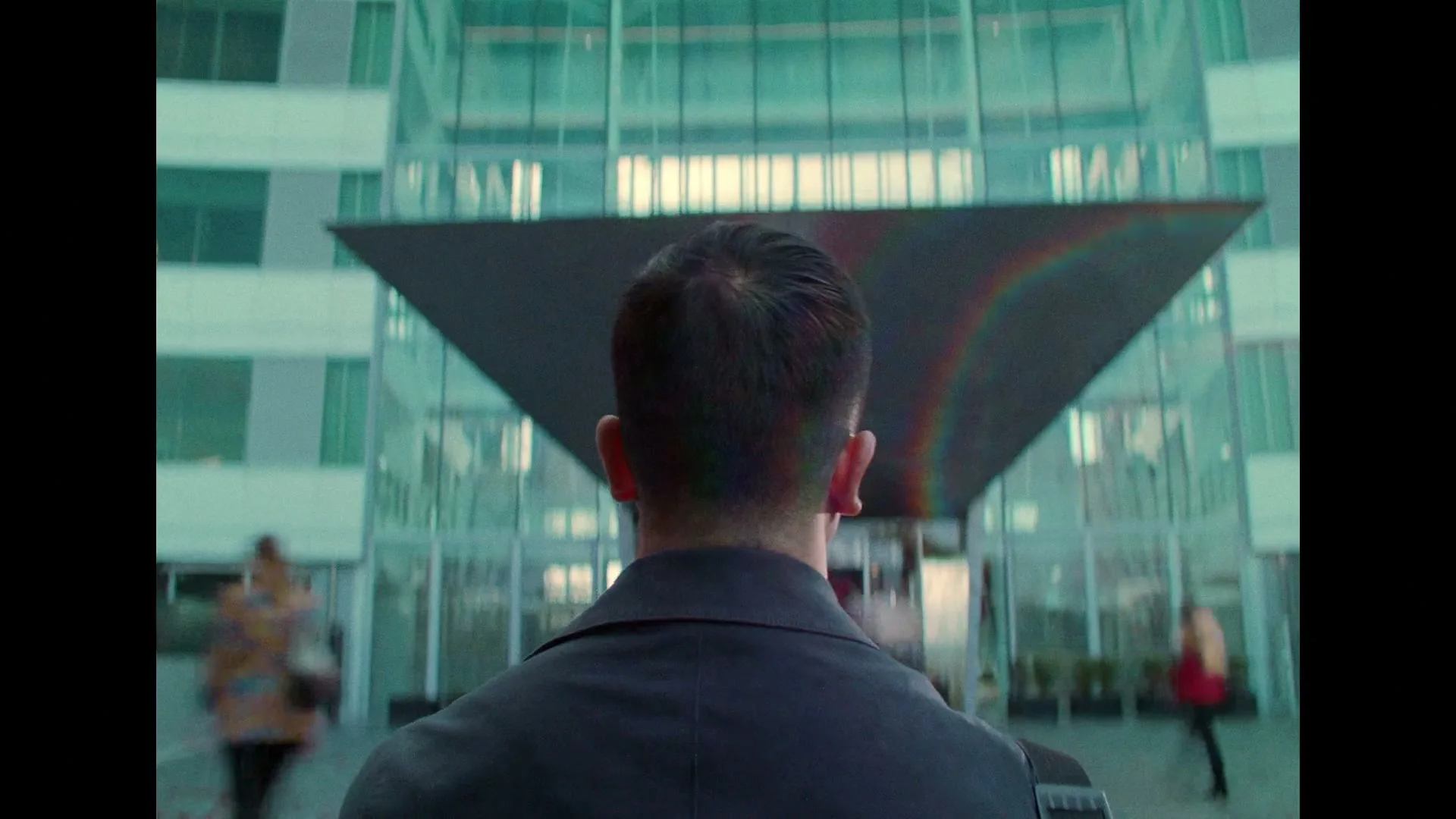 A man in a dark suit walks towards a modern glass building with a dark awning, seen from behind.