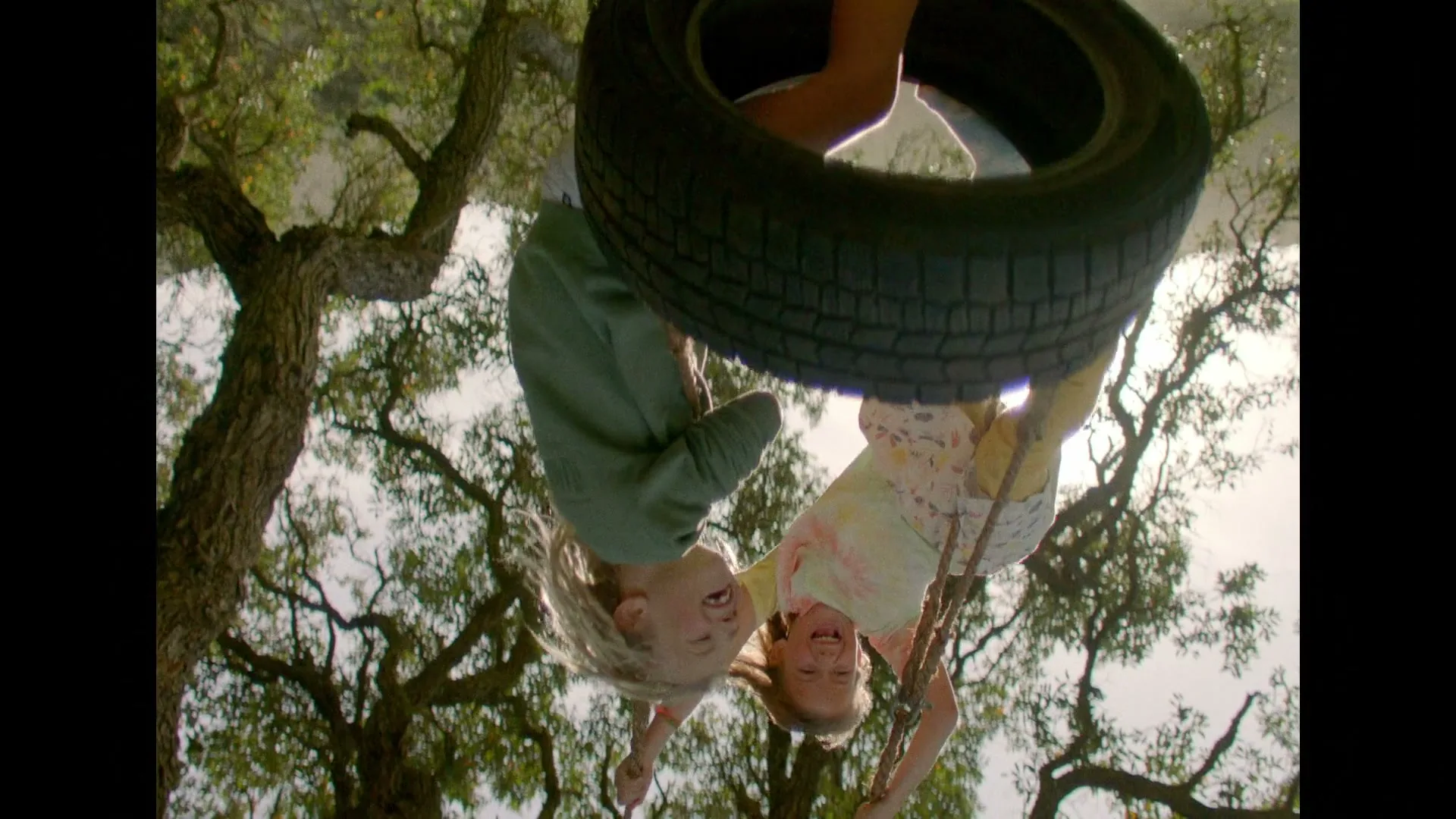 Two young children, a boy and a girl, hang upside down from a tire swing, laughing joyfully against a bright sky and green trees.