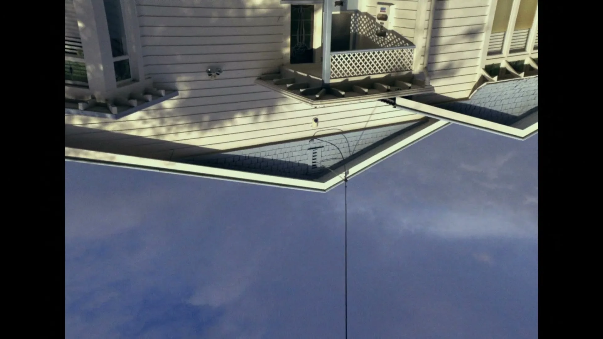 An upside-down shot of a white house with a blue roof and a small balcony, with a clear blue sky below it.