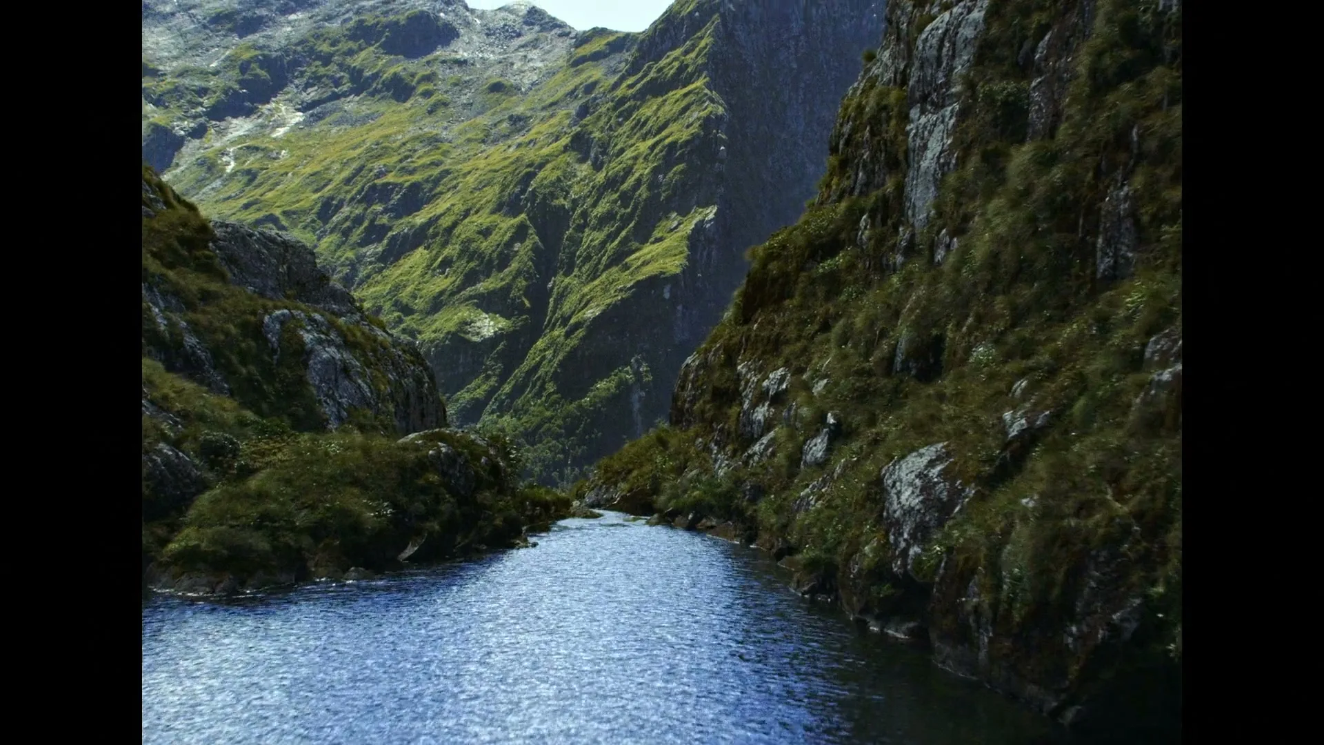 A wide shot of a river flowing through a valley with steep, green, rocky mountains on either side under a bright sky.