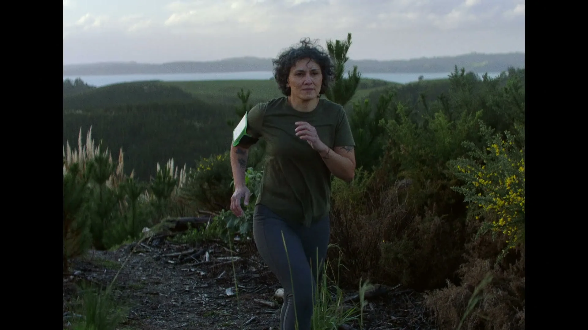A woman with curly hair, wearing a green t-shirt and grey leggings, runs on a dirt path through a hilly, forested landscape with a body of water in the background.
