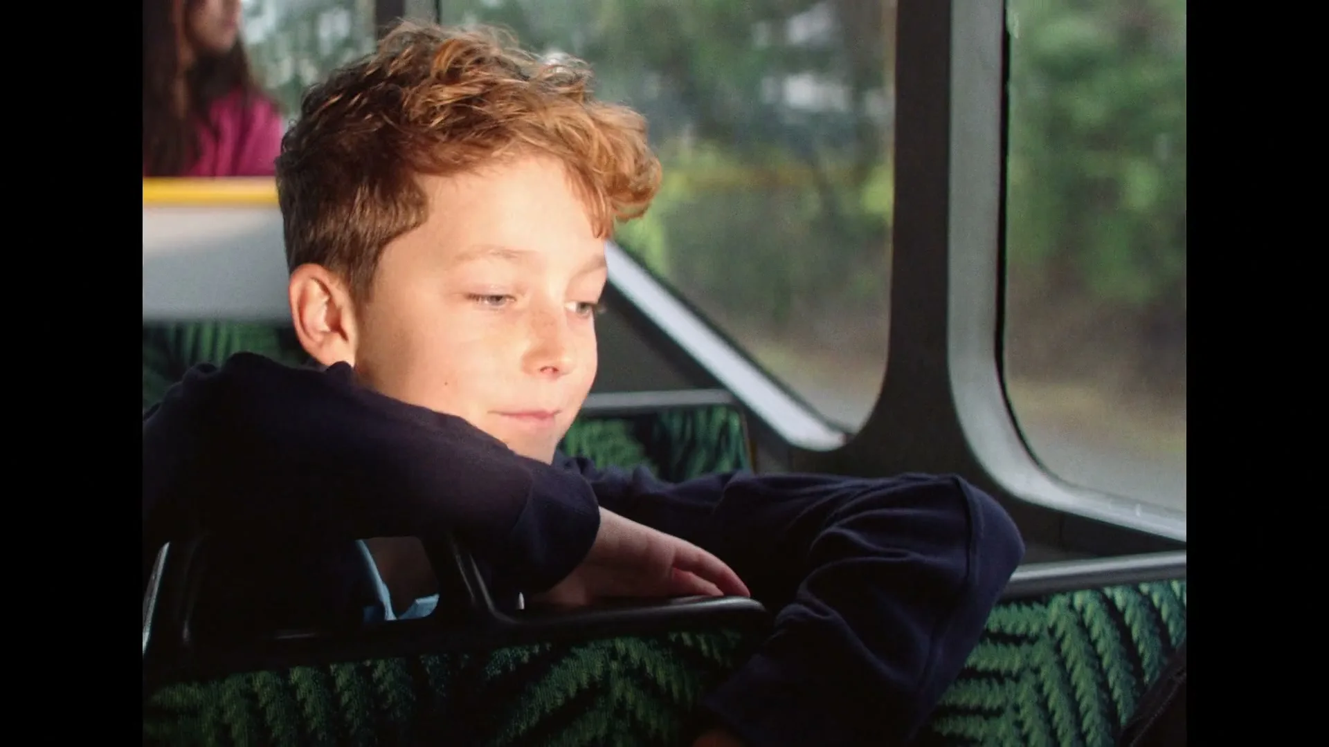 A young boy with curly hair sits on a bus, looking out the window and smiling, holding a phone.
