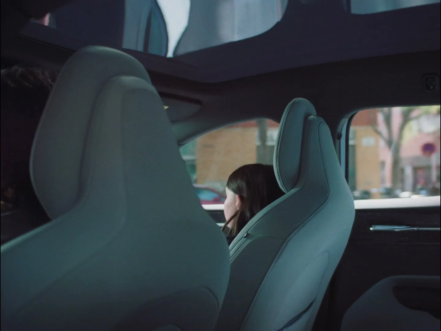 A young girl with dark hair sits in the back seat of a car, looking out the window with a thoughtful expression.
