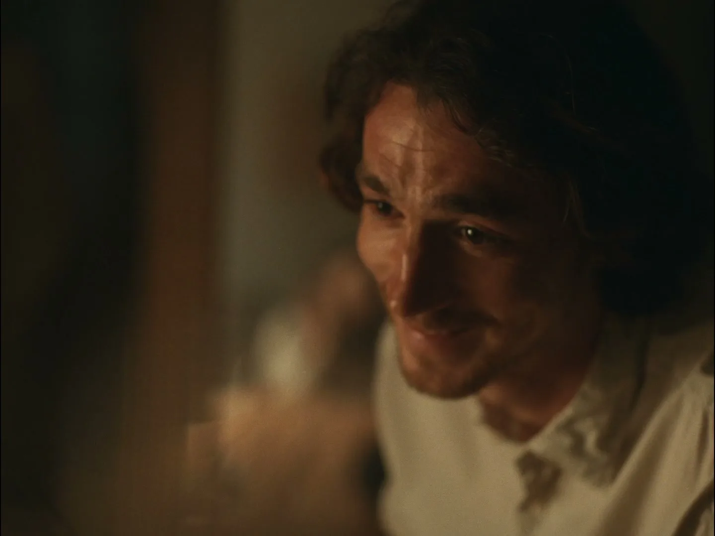 A man with curly brown hair and a white shirt looks down and smiles softly in a dimly lit interior setting.