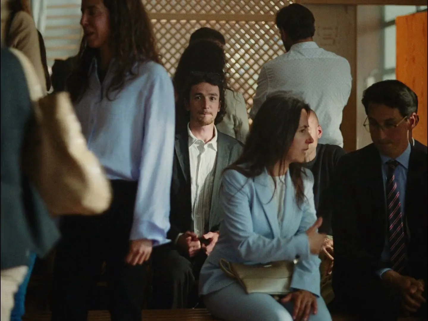 A diverse group of people, some seated and some standing, are gathered in a bright, modern interior space, with a man in a suit looking directly at the camera.
