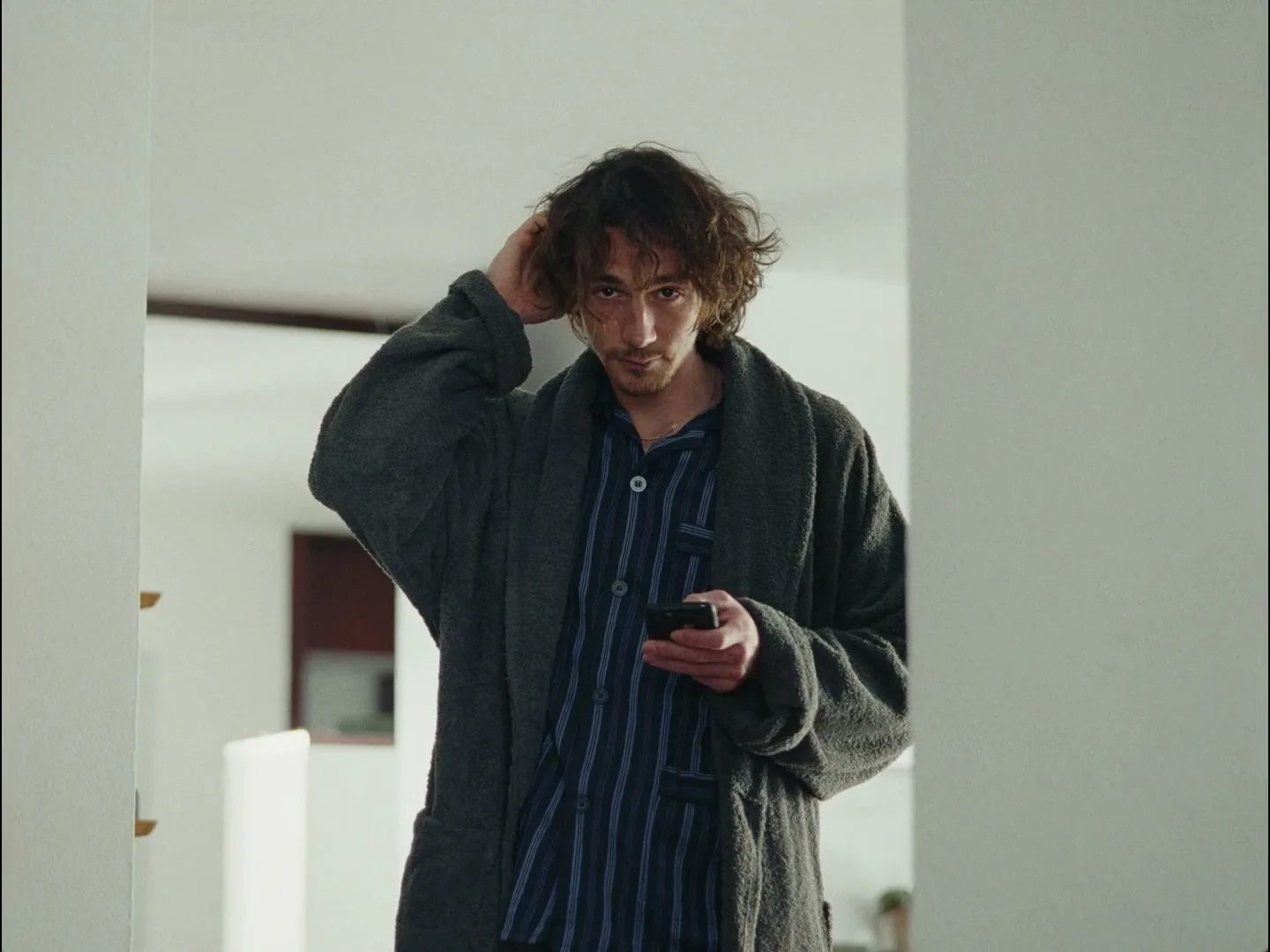 A young man with messy hair, wearing a bathrobe and pajamas, holds a phone and looks at the camera in a bright hallway.