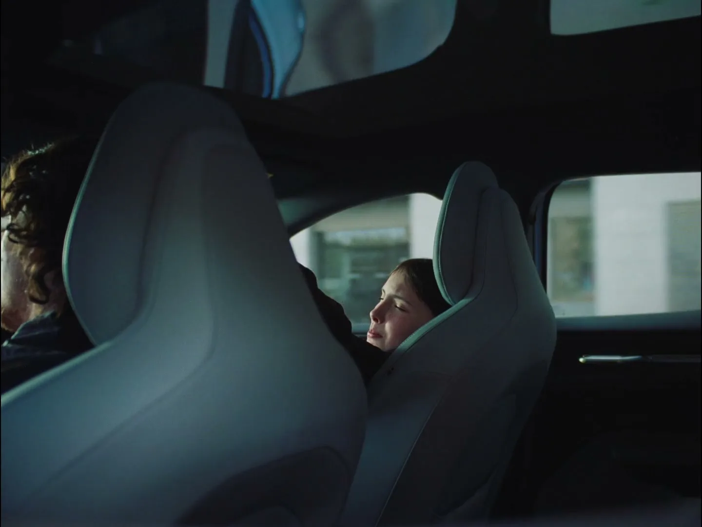 A young girl with dark hair, wearing a dark top, is seen from the back seat of a car, looking out the window with a slight smile.