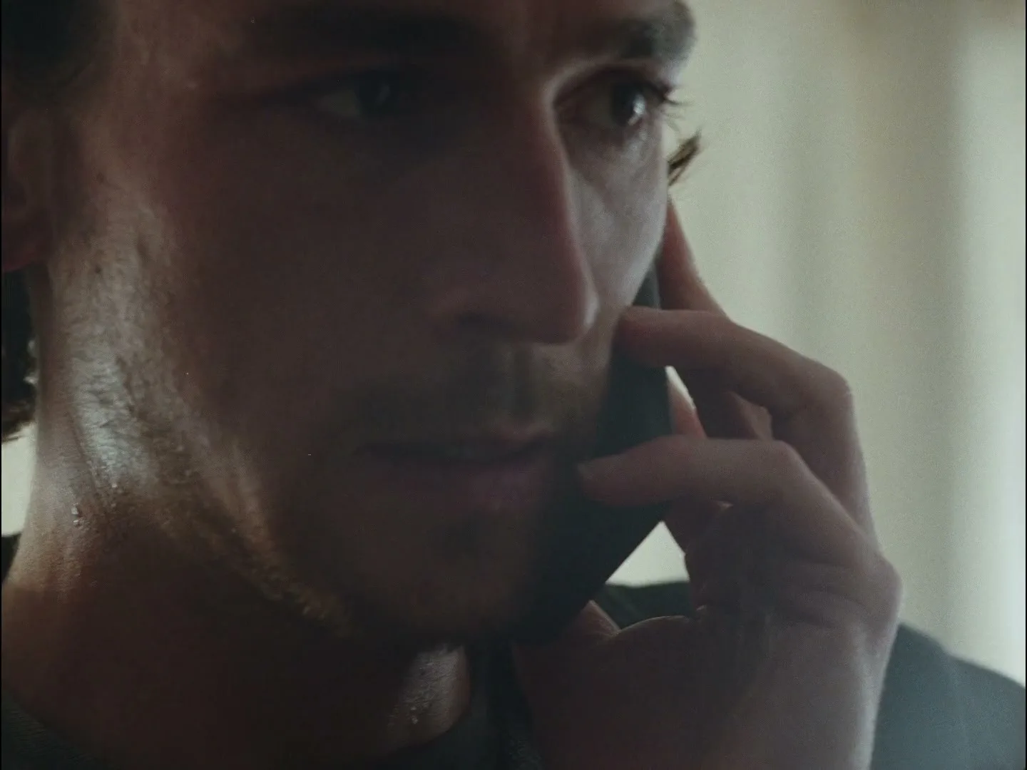 A man with dark, curly hair and stubble holds a phone to his ear, listening intently in a softly lit room.