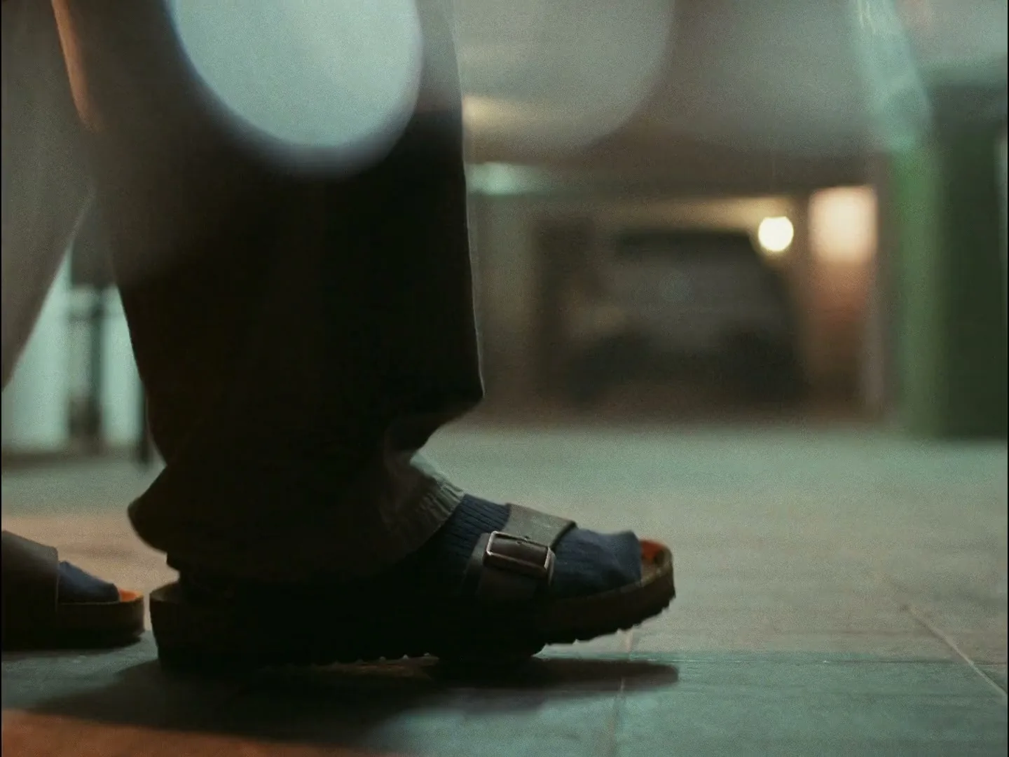 A person's foot in a sandal and sock steps on an aluminum can on a tiled floor in a dimly lit interior.