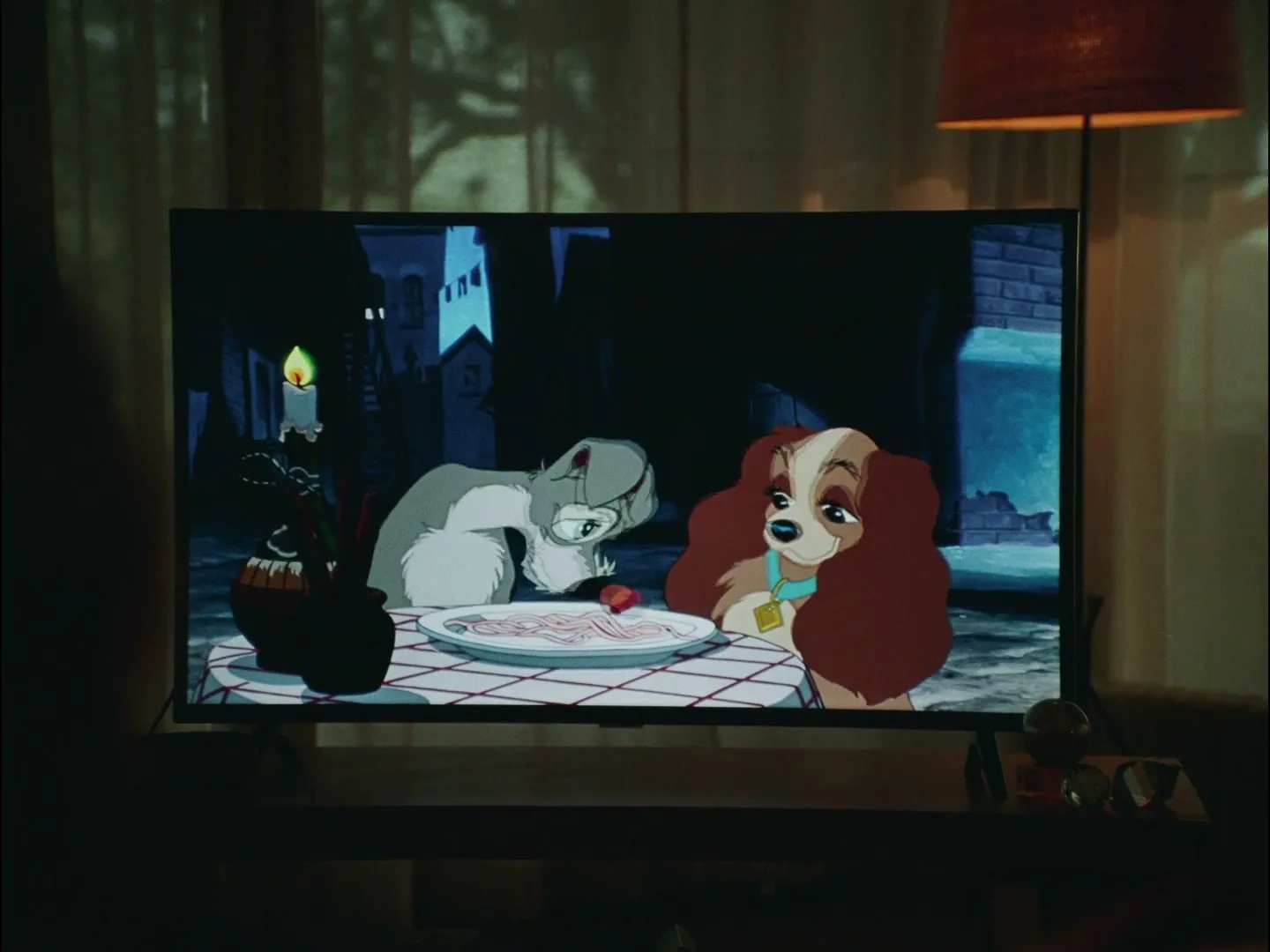 A television screen shows an animated scene of two dogs, Lady and the Tramp, sharing spaghetti at a candlelit table in a dark alley.