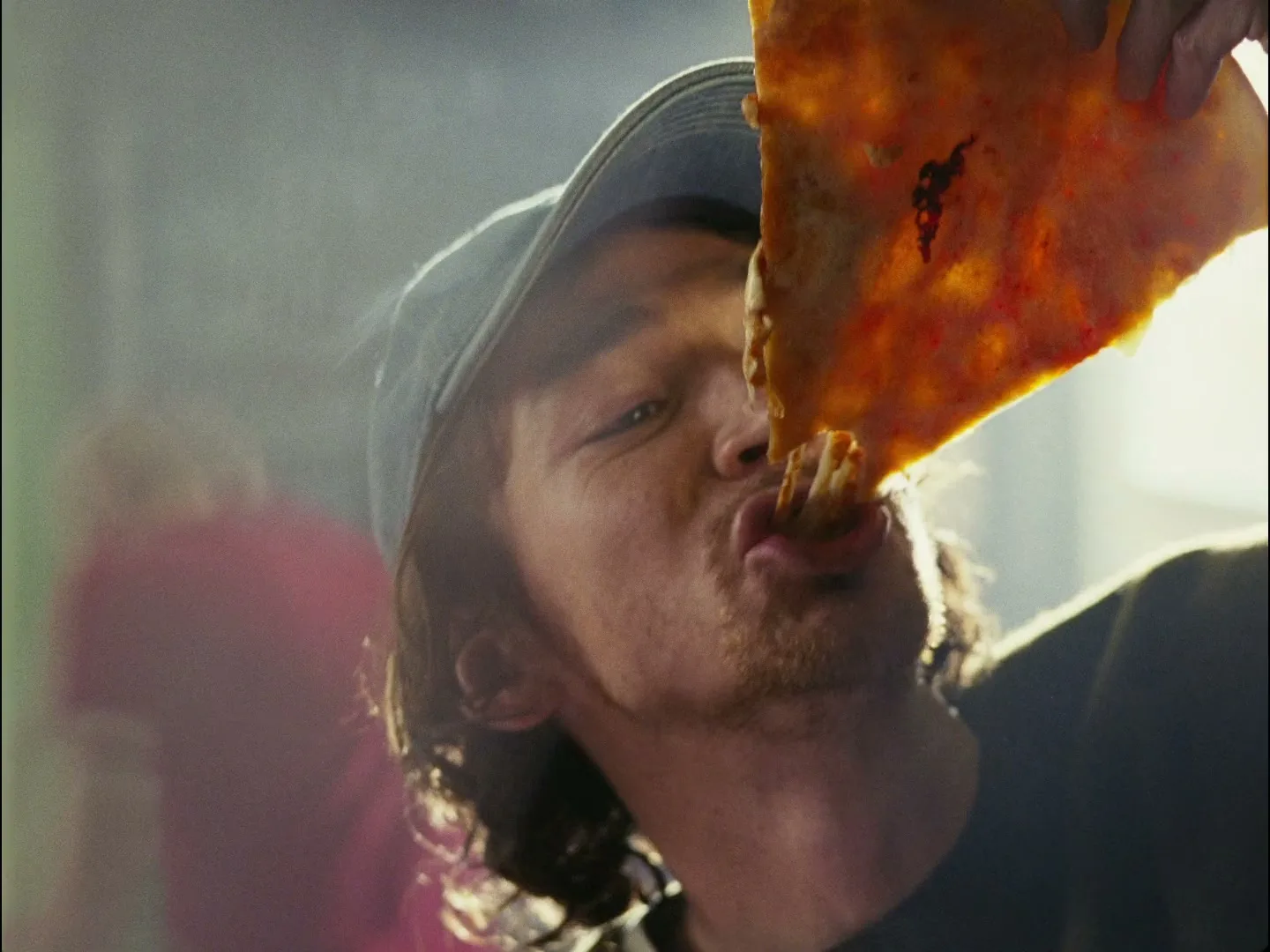 A young man with long hair and a baseball cap eats a slice of pizza, pulling a long string of cheese from his mouth, in a bright, hazy interior.