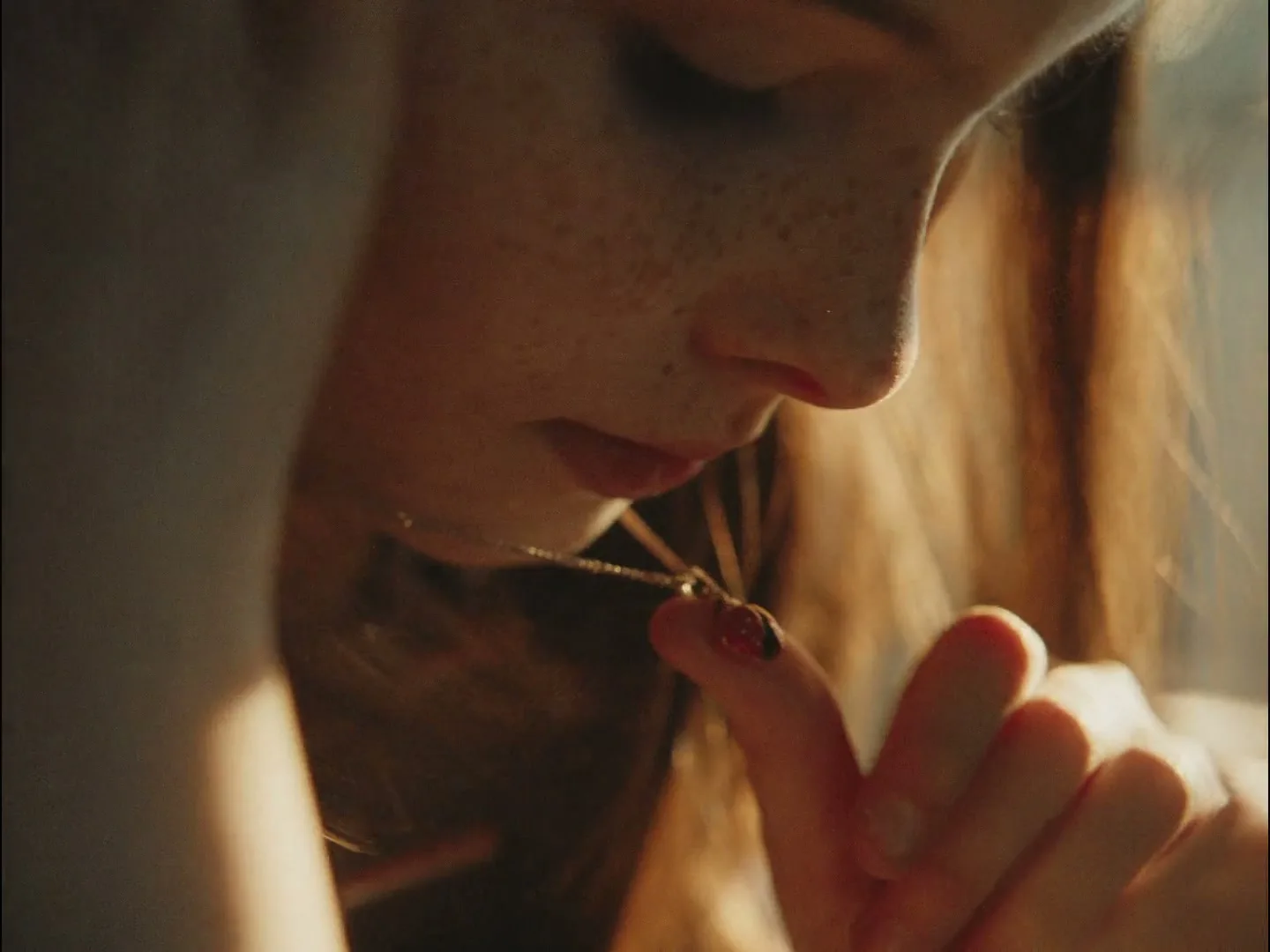 A young woman with freckles looks down at a small ladybug charm on a necklace, holding it gently with her fingers.