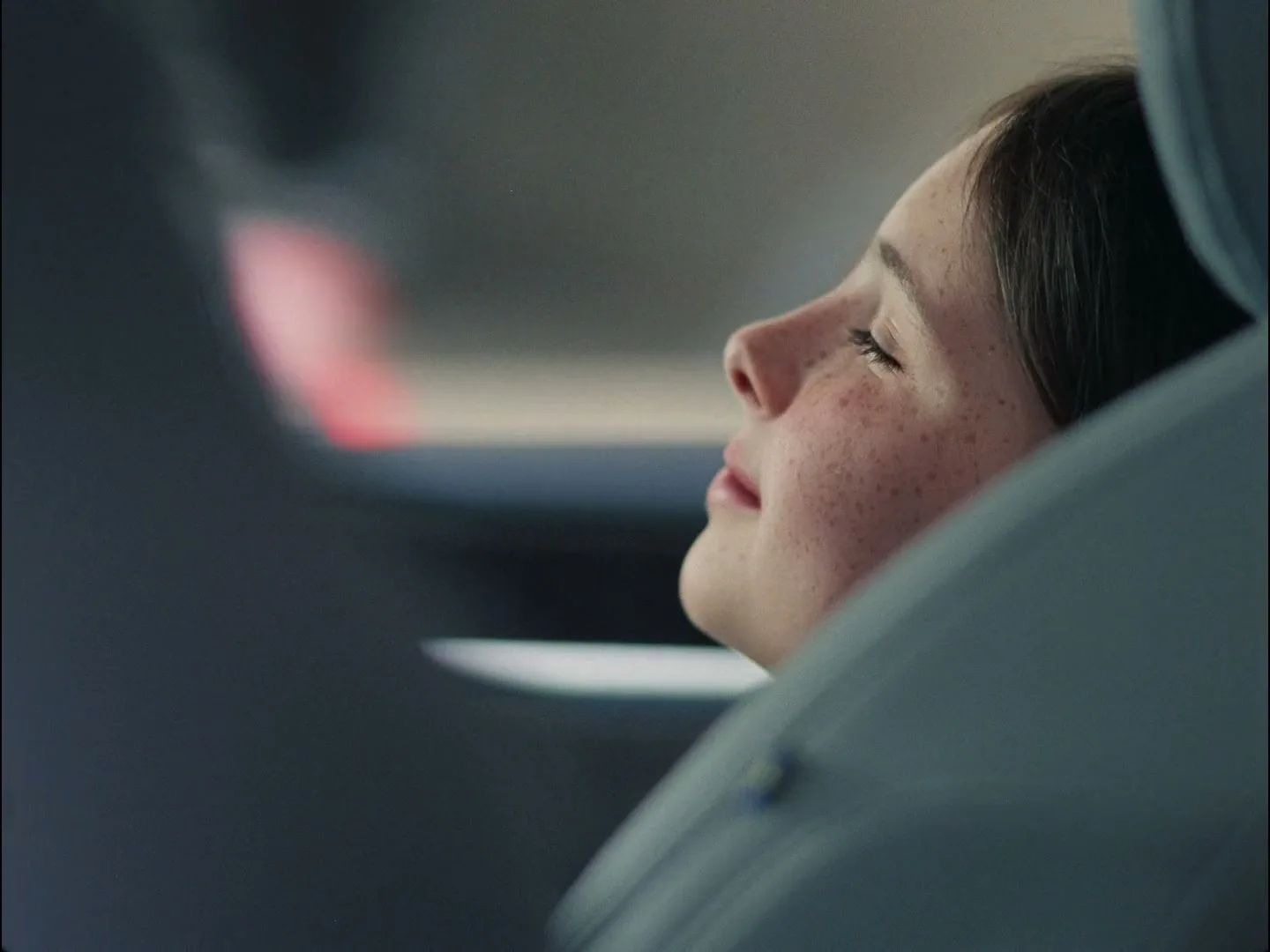 A young girl with freckles rests her head against a car seat, eyes closed, as the car moves, seen from a medium closeup.