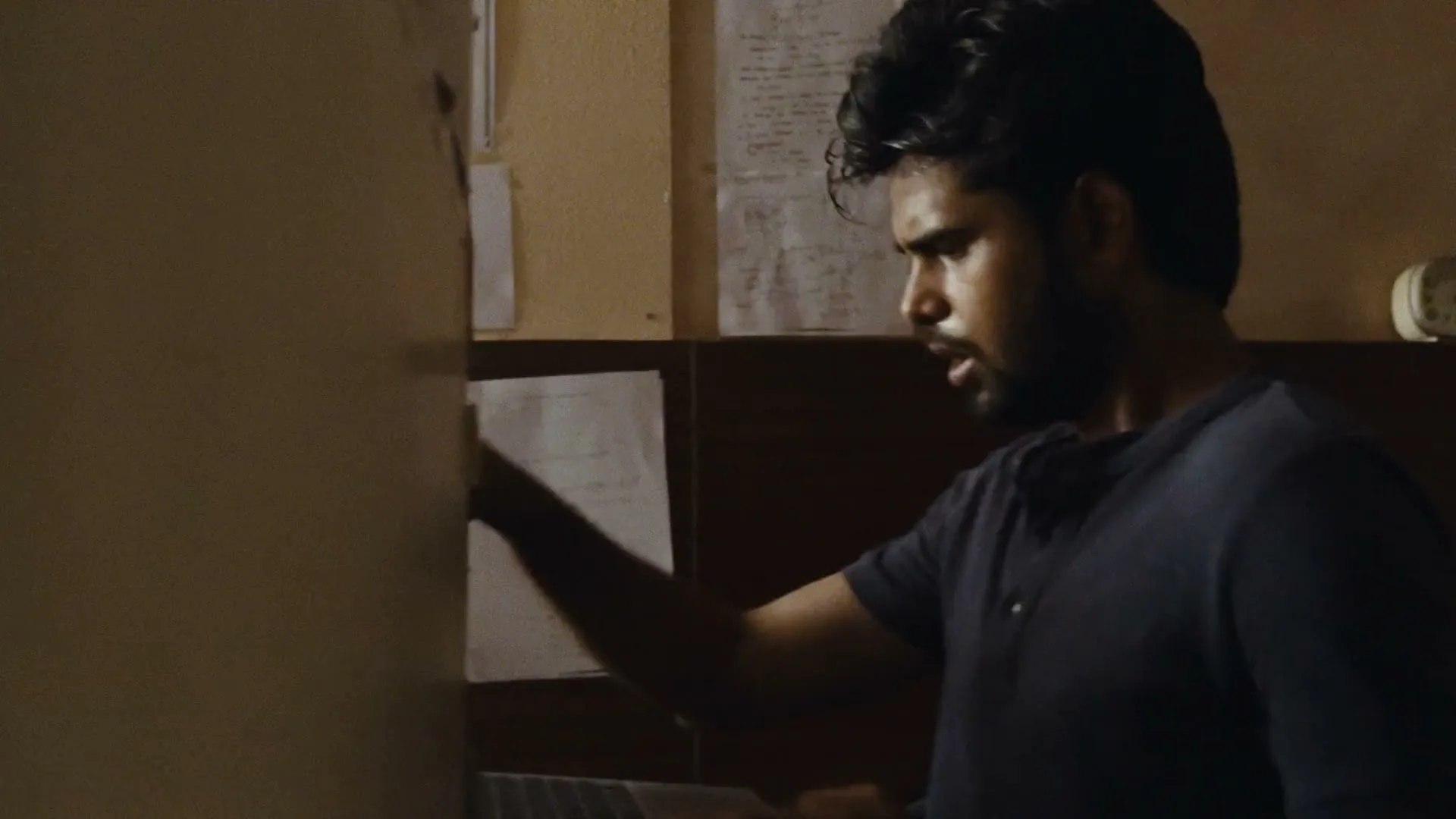 A young man with dark hair and a beard, wearing a dark t-shirt, works on a laptop in a dimly lit room.