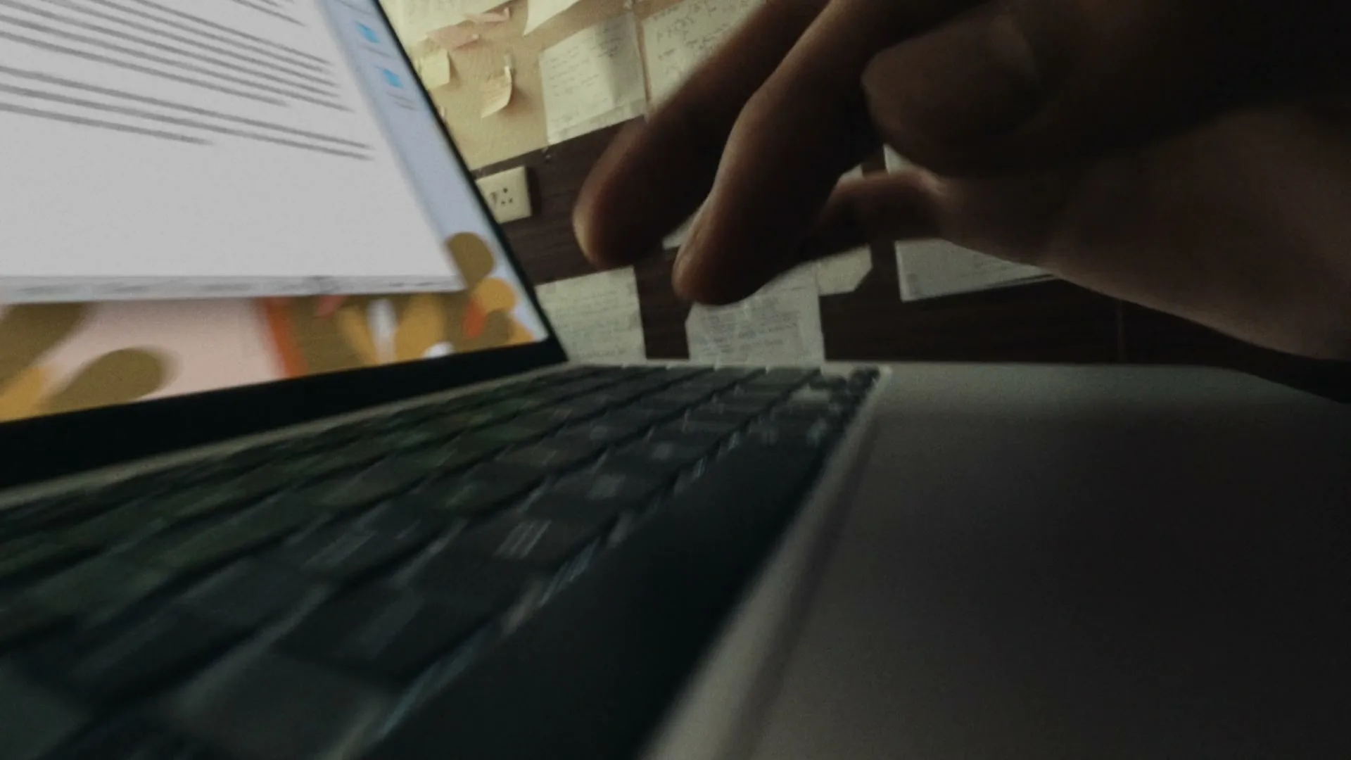A person's hand types on a laptop keyboard, with a wall covered in sticky notes and papers in the background.