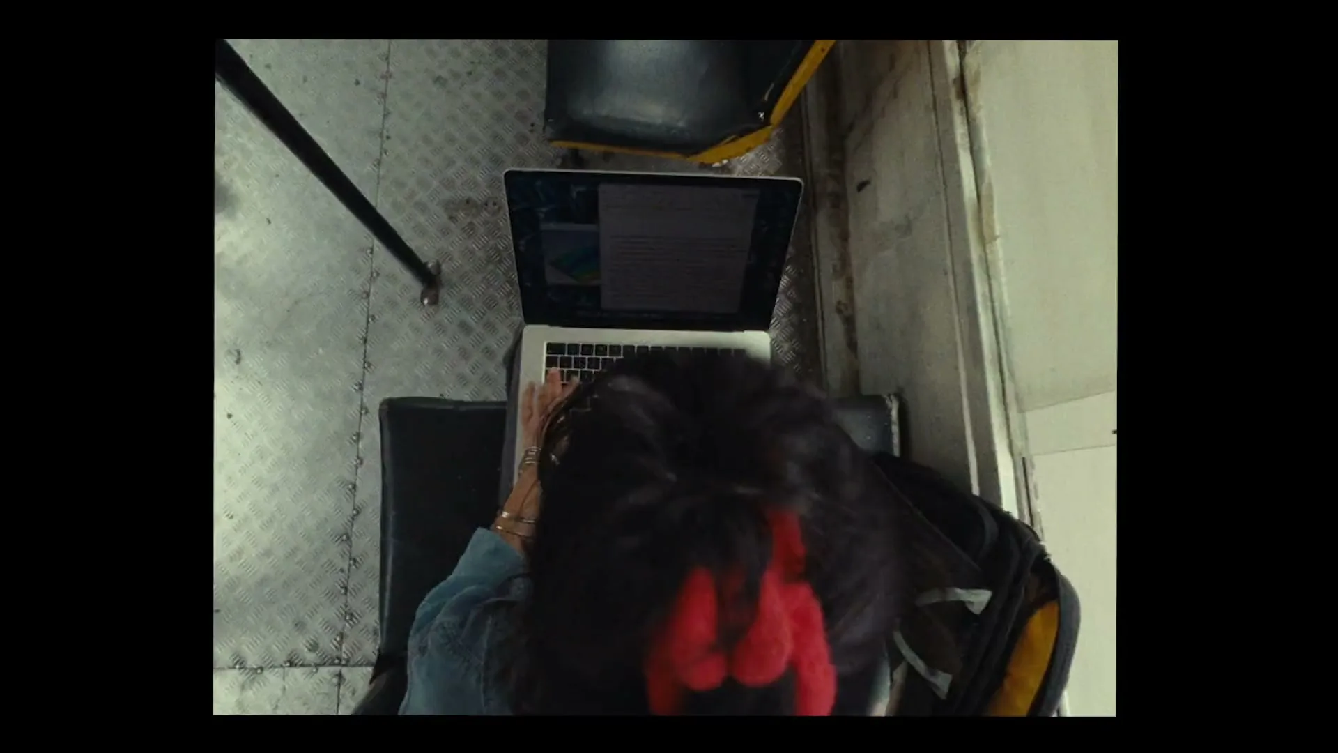 A person with dark hair tied with a red scrunchie types on a MacBook laptop while sitting on a bus, seen from an overhead perspective.